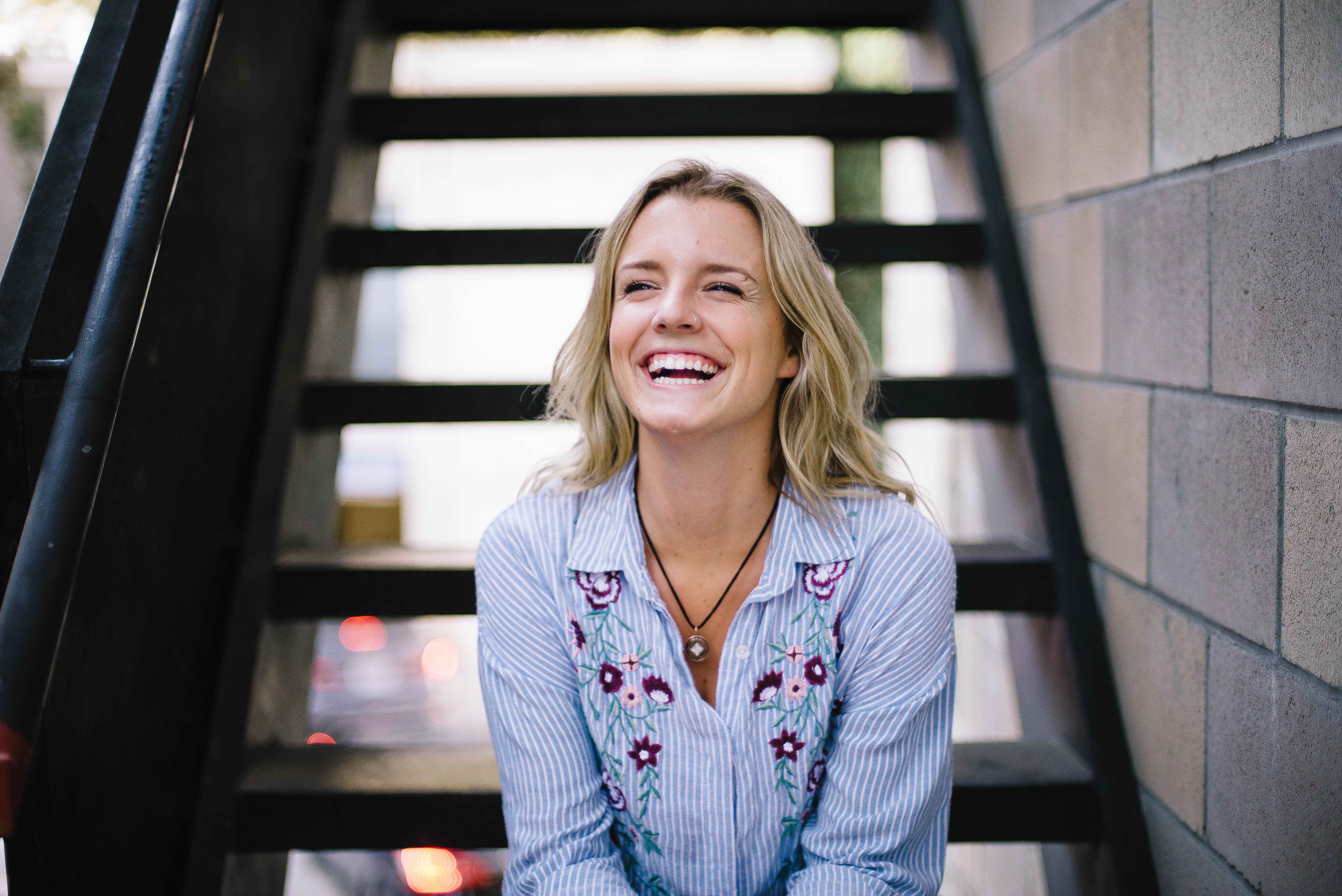 A smiling person with blonde hair sits on outdoor stairs, wearing a striped shirt with floral embroidery.