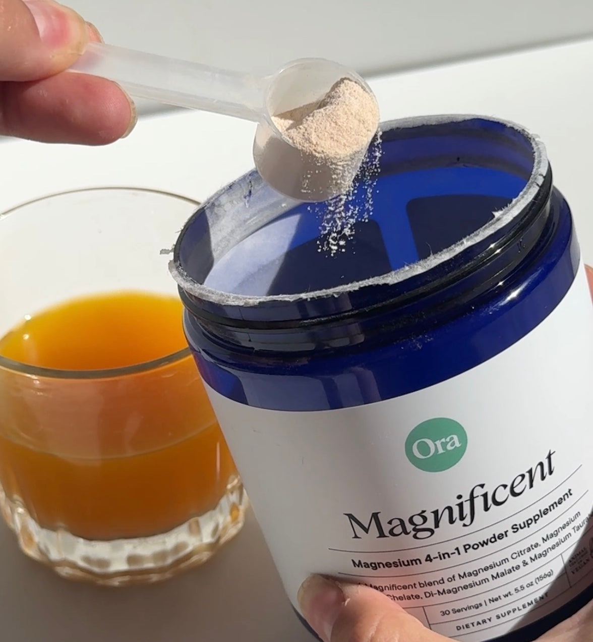 A scoop of powdered supplement is being added to a jar labeled 'Magnificent' next to a glass of orange juice.