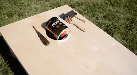 Unfinished cornhole board with a can of poly and brushes sitting on top of it
