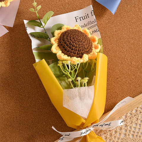 Sentence with new product name: A handmade crochet flower with a brown center and yellow petals, attached to the Small Bouquet of Crochet flowers with greenery and a yellow wrapper, placed on a cork background.