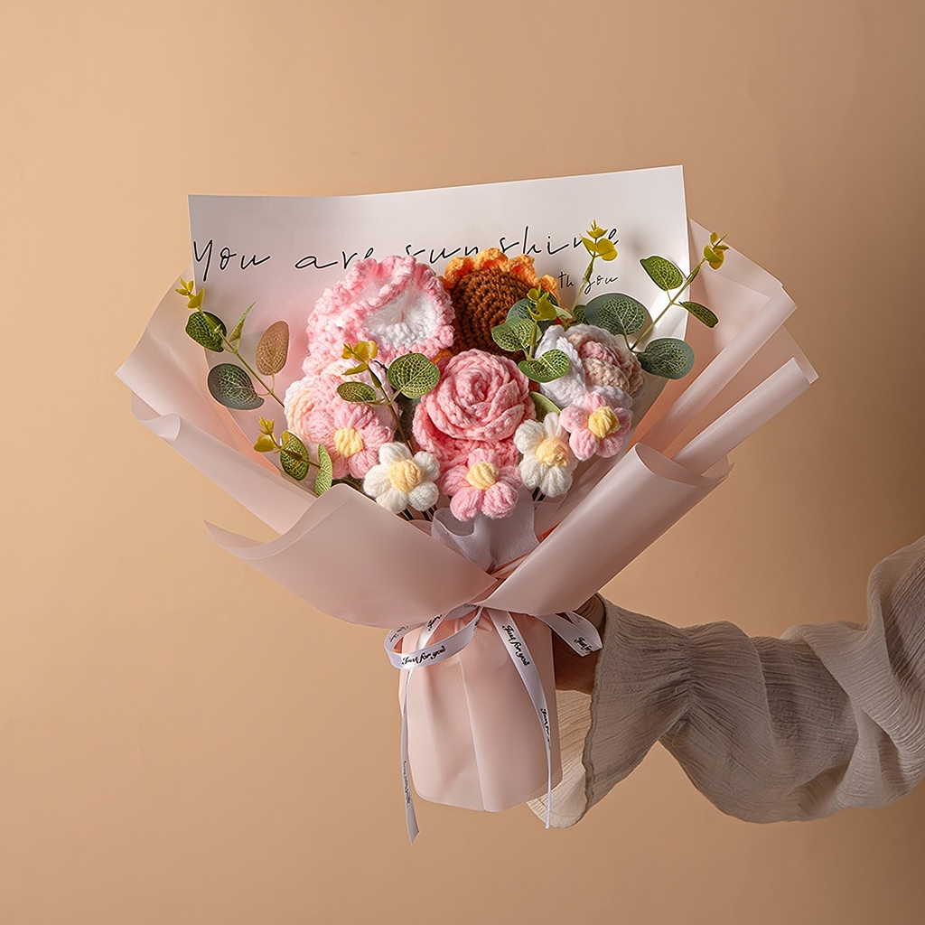 A hand holds the Crochet Sunflower Bouquet: Blooms That Never Wilt, featuring pink, white, and yellow flowers in pale pink paper. A note says, "You are my sunshine," adding handmade charm and lasting beauty to this heartfelt gift.
