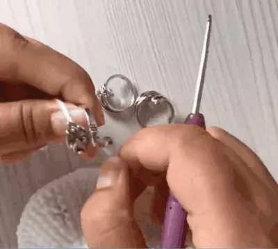 Hands crocheting with a purple hook and white yarn, featuring two rings, including the adjustable Yarn Tension Control Ring, alongside an open locket resting on a white surface.