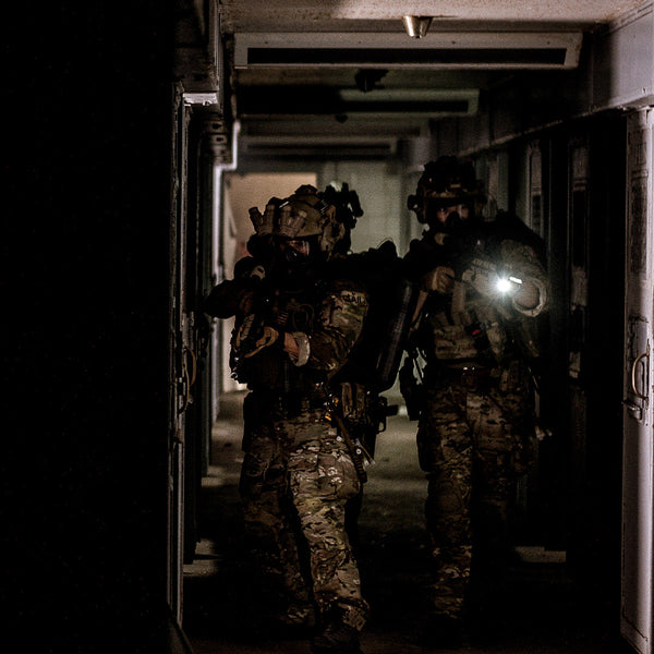 Men wearing MultiCam uniforms, plate carriers and wielding rifles scan a dark hallway.