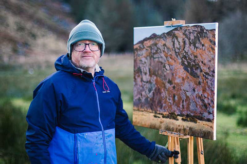 Photo of artist Kieran Guckian standing in front of a painting of a craggy mountain on an easel on location in Lough Dan Wicklow