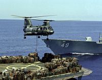 A Helicopter Combat Support Squadron 5 (HC-5) CH-46 Sea Knight helicopter tansports cargo from the helicopter pad of the combat stores ship USS SAN JOSE (AFS-7) during a vertical replenishment operation. The bow of the guided missile cruiser USS VINCENNES (CG-49) is in the background. - 1986