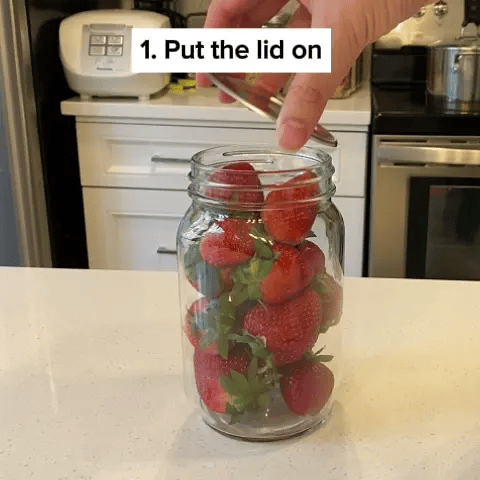 A hand placing a lid on a jar full of strawberries.