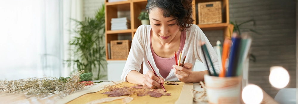 A woman of Asian descent is wearing a white cardigan and coloring at her kitchen table. 