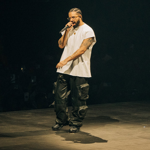 Drake stands center stage with a microphone in hand, performing before an audience. He sports a modern street style look with a plain white tee and unique black cargo pants adorned with multiple pouches. His hairstyle features cornrows that extend into braids, complemented by a well-groomed beard. The lighting highlights him against the dark backdrop of the venue, emphasizing the live performance atmosphere.