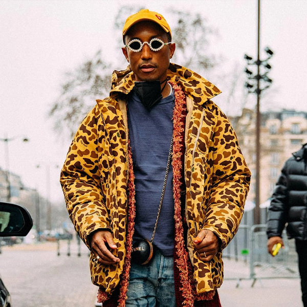 Pharrell Williams poses for a photo on a city street, making a fashion statement with his unique ensemble. He wears a bold leopard print jacket with a furry collar and a dark navy turtleneck underneath. His accessories include a yellow baseball cap, possibly worn backward, and distinctive sunglasses adorned with pearl-like embellishments around the rims. A black face mask is visible under his chin, indicating a setting during or post-pandemic times. He also sports a chain with a pendant, and the word "CHANEL" is discernible on one of his accessories, hinting at designer fashion elements in his outfit.