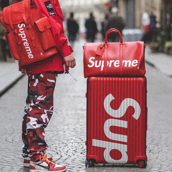 A person clad in red Supreme streetwear, including a branded jacket, camo pants, and sneakers, stands next to a matching red suitcase and carries a red shoulder bag, all adorned with the same bold white logo.