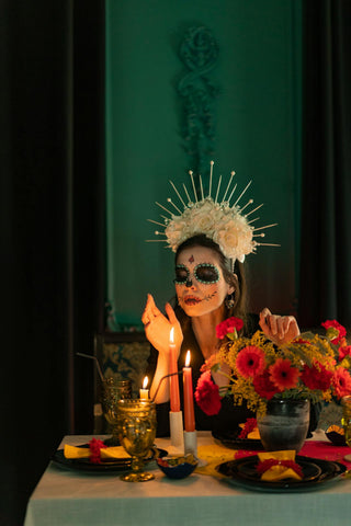 A woman in dia de los muertos make up enjoying a candle lit dinner. There is a bouquet of red flowers on the table.