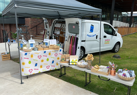 Outdoor pop-up shop out of the back of a small, white cargo van parked half on a grassy area and half on the sidewalk in front of a tall building. Under a large gray tent are two tables filled with dispensers and baskets of sustainable cleaning and personal care products.