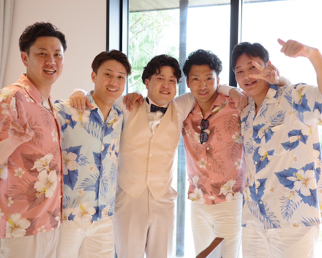 リゾート結婚式で新郎と友人男性ゲストが肩を組んで記念撮影。新郎はベージュのタキシード、ゲストはピンクやブルーの花柄アロハシャツをお揃いで着こなし、南国らしい華やかな雰囲気を演出。