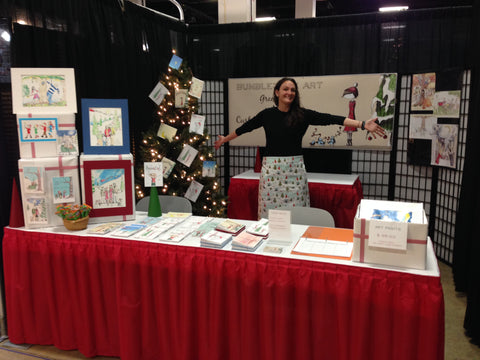 A woman standing behind a craft show booth display of cards and artwork