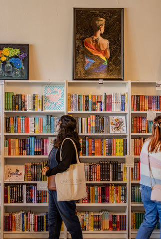 two femme people walk by a colorful bookshelf, slightly in motion. there is a painting of a person with short hair and a butterfly tattoo between their shoulder blades wearing a pride flag draped over them.
