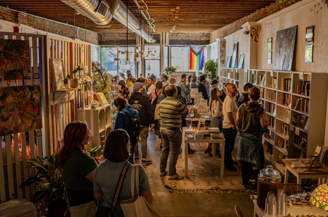 a bookstore full of people with shelves lining the right side of the room. a pride flag is visible hanging from the window in the back of the picture