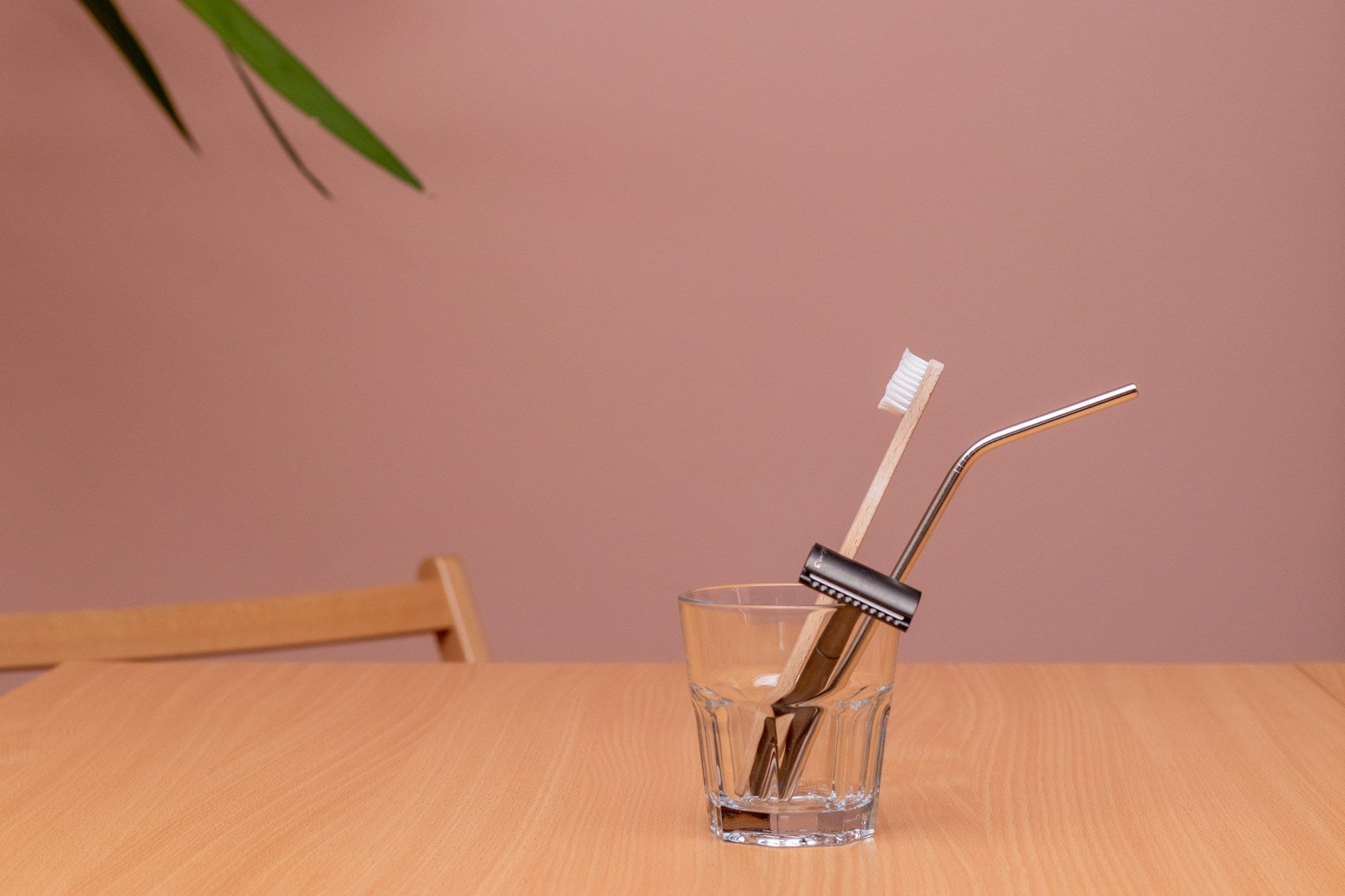 A clear glass cup holding a safety razor, bamboo toothbrush, and stainless steel straw in front of a mauve background