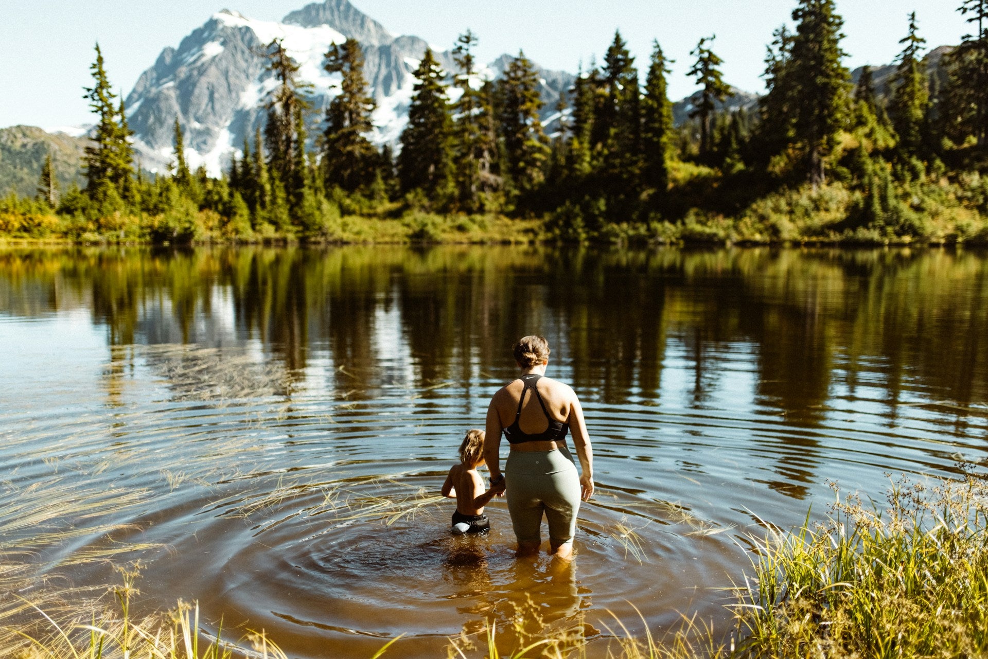 A woman and a young child walking into an alpine lake in the summer with gorgeous mountains in the background