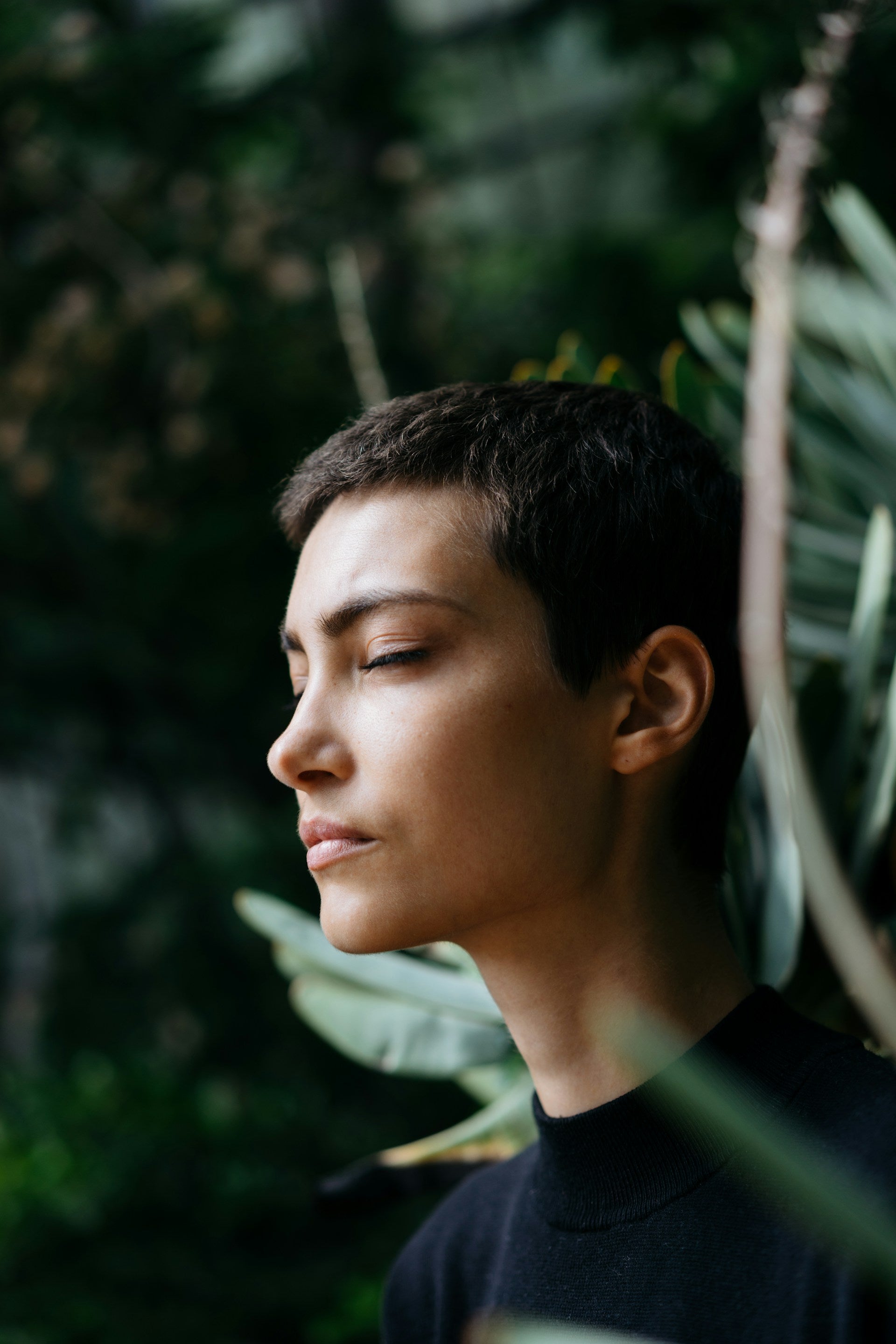 A short haired person closing their eyes in meditation with greenery in the background
