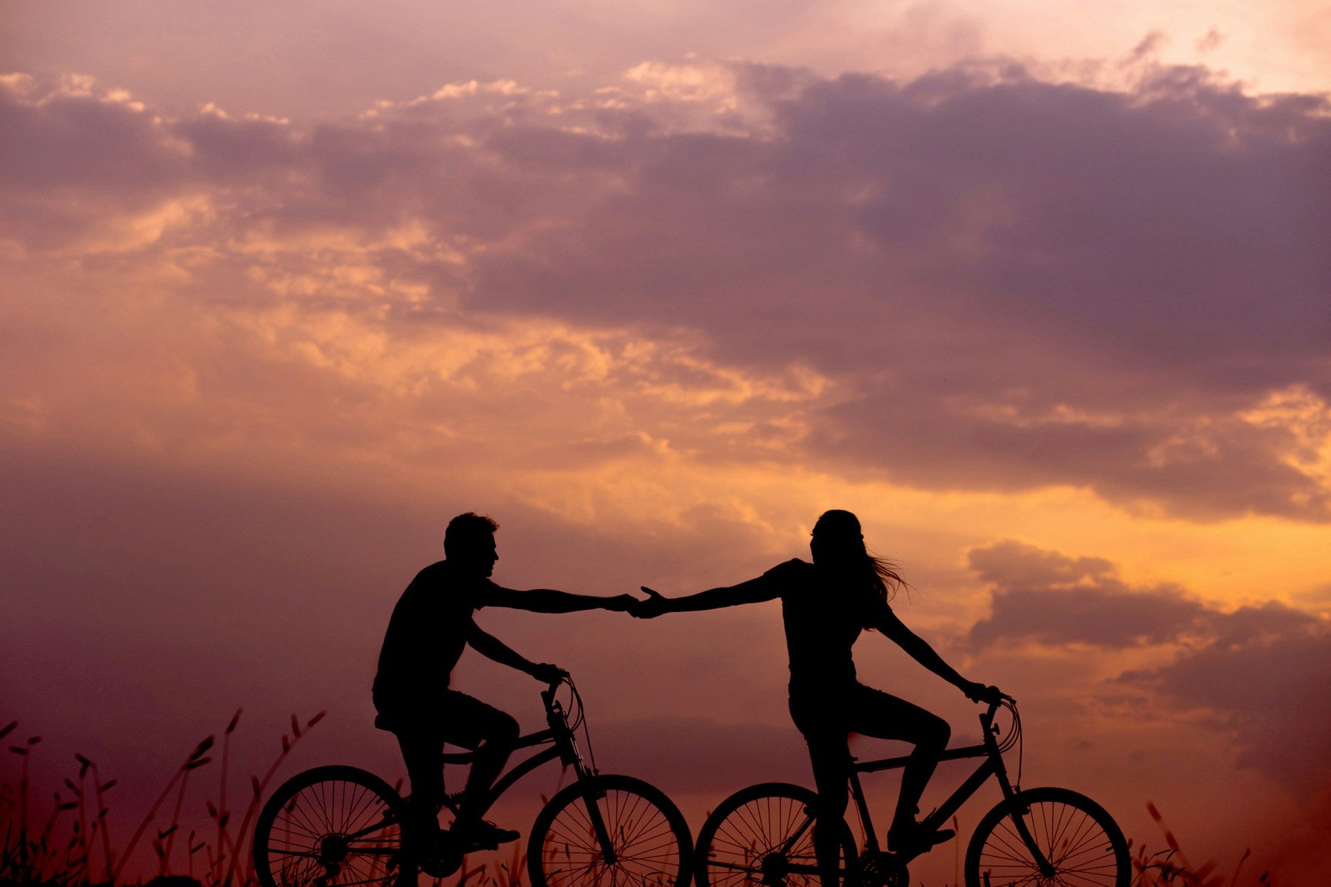 A couple riding bikes while reaching for each other with a pink sunset sky in the background