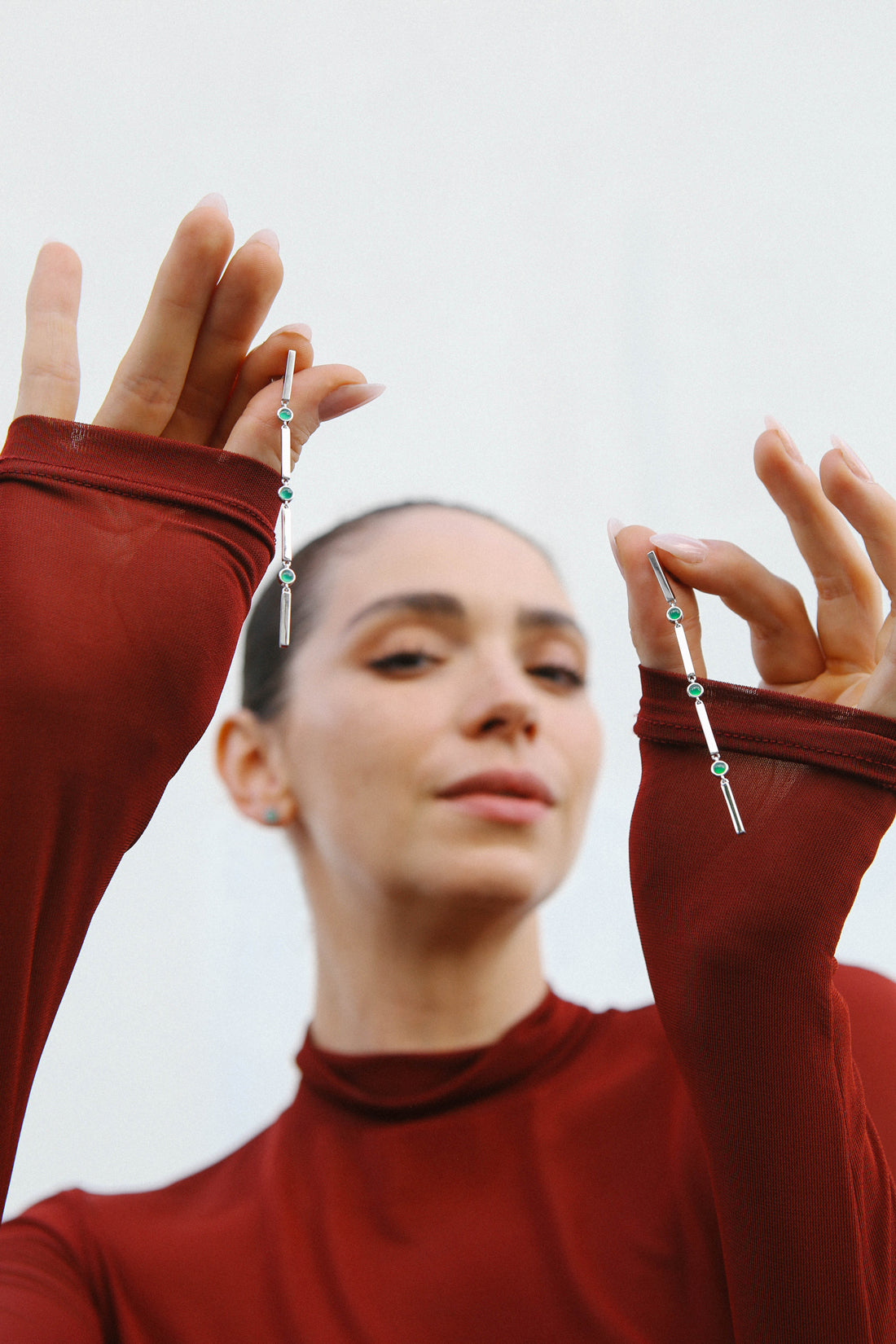 Model wears a red long sleeve top, and holds up a pair of silver earrings featuring three green stones.