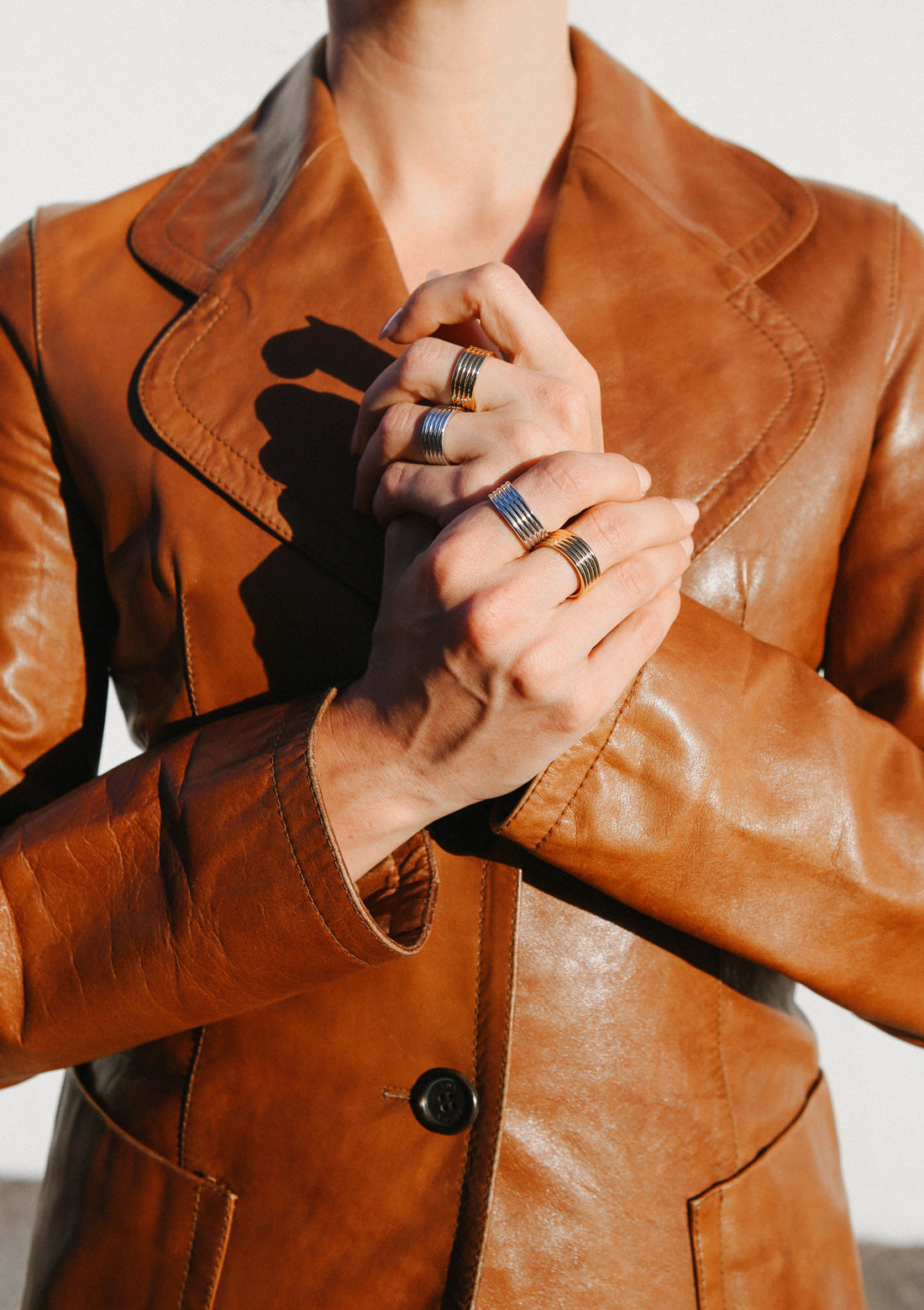 Model wears a tan brown leather blazer jacket, with her hands clasped together, showcasing four gold and silver rings.