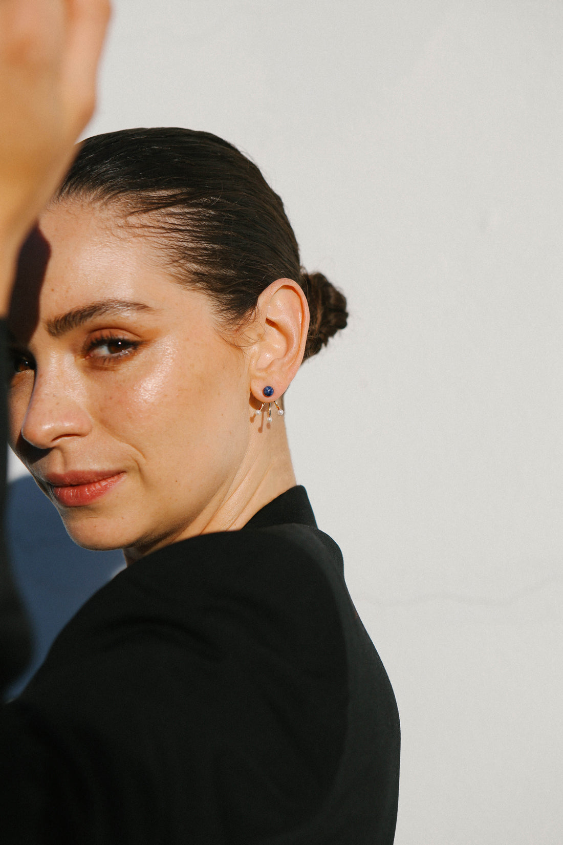 Model wears a black jacket and holds one arm up, which casts a slight shadow on her face. The feature is a single earring of three pearls and one blue stone stud.