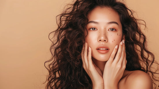 Beautiful asian girl with long wavy hair. Her fingertips are placed on her cheeks in front of a beige background.