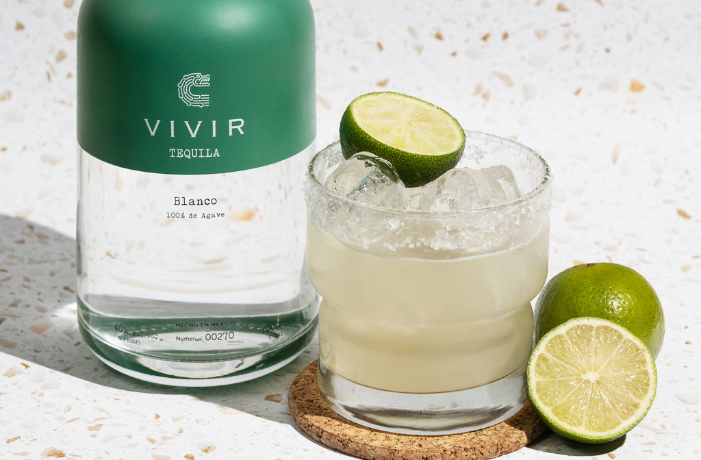 A bottle of VIVIR Tequila Blanco positioned next to a Margarita cocktail and some limes.