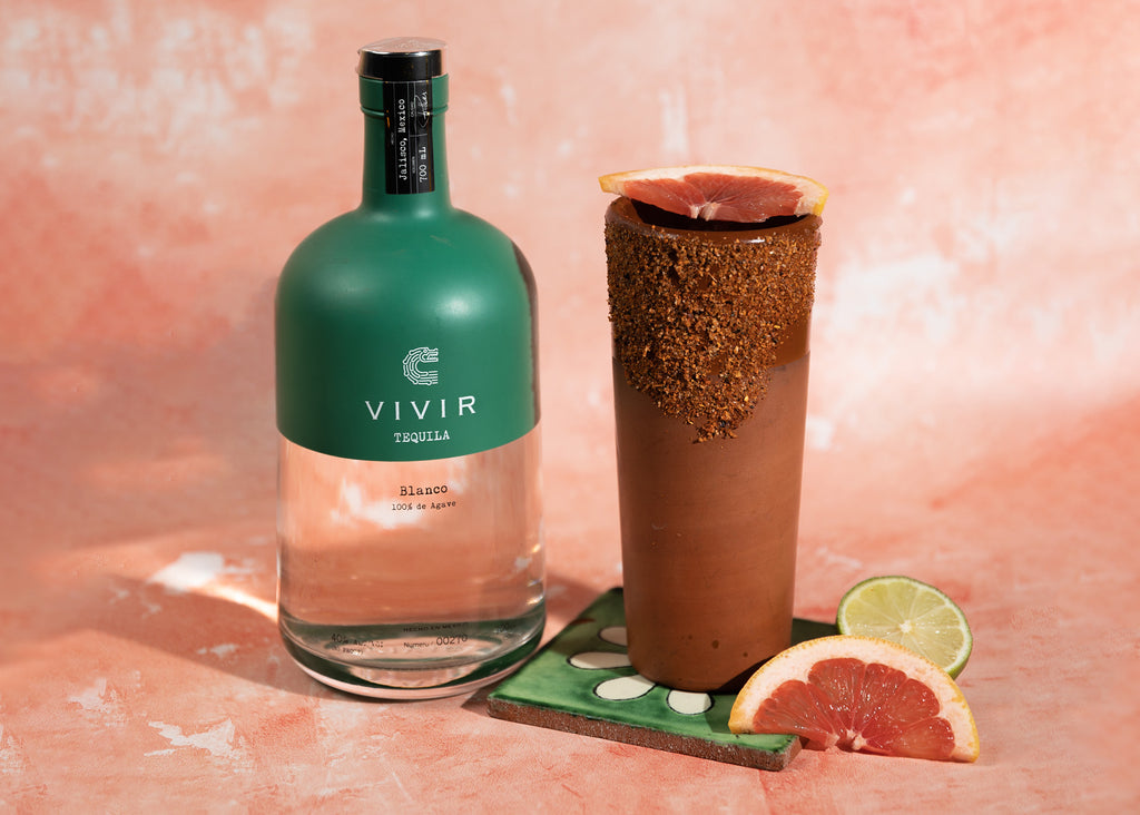 A bottle of VIVIR Tequila Blanco is positioned next to a Paloma cocktail with pink grapefruit wedges in front.