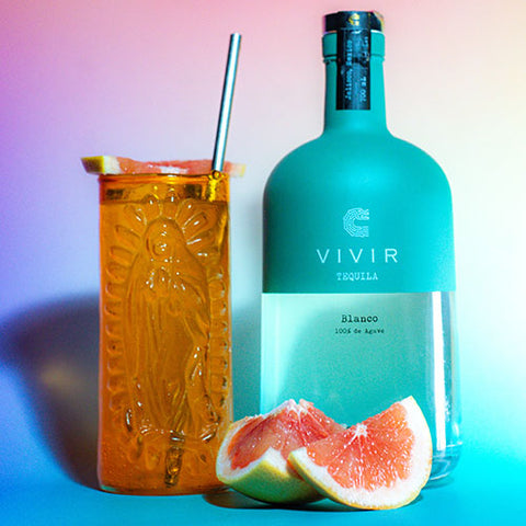 A Paloma cocktail is positioned next to a bottle of VIVIR Tequila Blanco with some pink grapefruit wedges in front.
