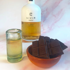 A bottle of VIVIR Tequila Reposado is positioned next to a sipping glass filled with Tequila and a pot of dark chocolate.