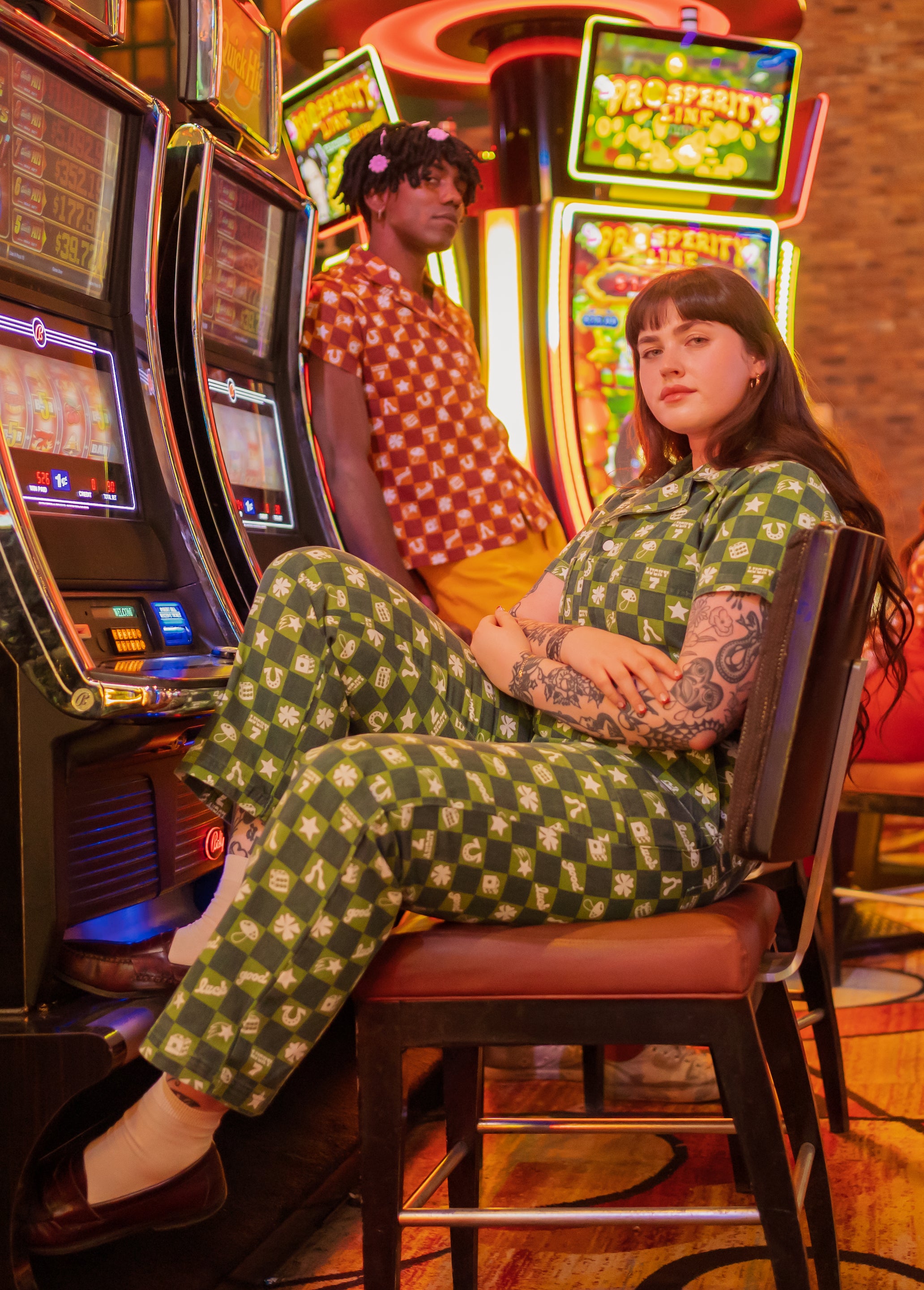 Sydney in Lucky Short Sleeve Jumpsuit in Green sitting in front of slot machine with Jerrod in the background wearing Pantry Button-Up in Spicy Mustard and Western Pants in Sunshine Yellow.