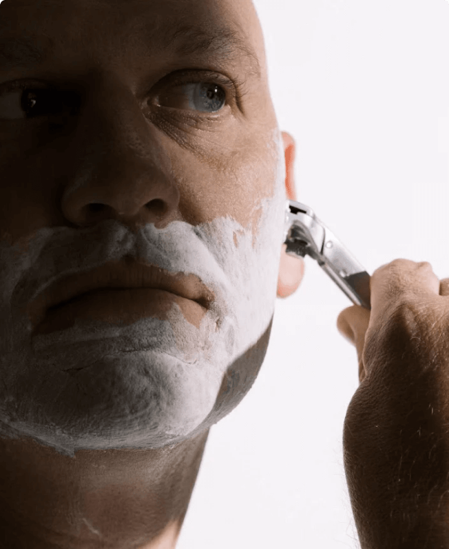 A man shaving his face with a safety razor, applying shaving cream.