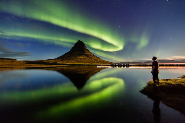 Vincent Ledvina standing under the aurora in Iceland with reflections on the water with a mountain in the background