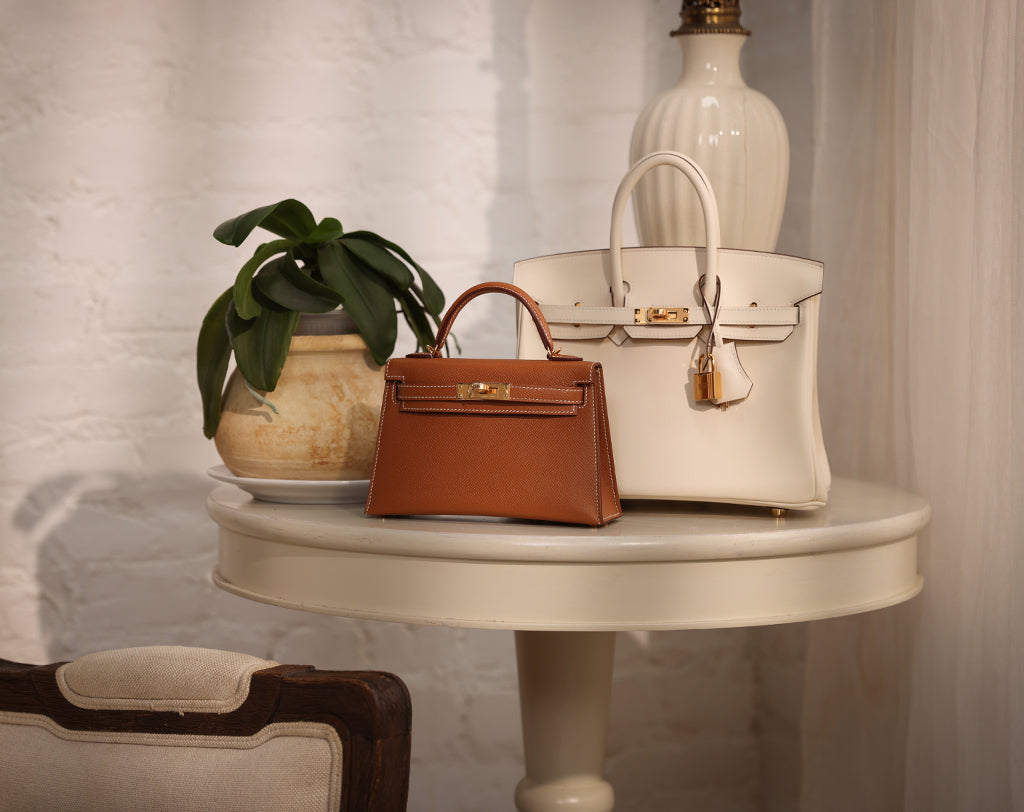 Still life image of an Hermès Gold Kelly 20cm and Nata Birkin 25cm on a table.
