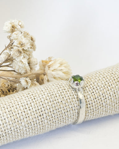 A silver stacking Ring set with a 5mm intensely green peridot that is supported with a linen wrap and cream coloured dried flowers in the background