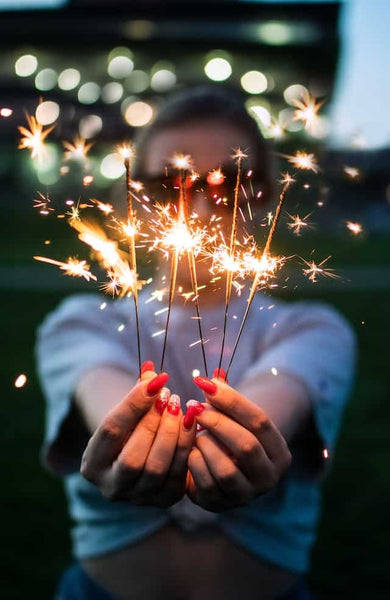 A person holding sparklers with a vivid display of sparks, showcasing red handcrafted false nails, with a blurred background lighting up the twilight