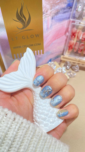 A hand presents a set of sparkling blue false nails with delicate gold accents, complemented by a mermaid tail decoration and the LTGlow branding in the background