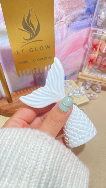 A close-up of a hand showcasing a pastel blue acrylic nail, complemented by a white mermaid tail decoration, with the golden LT GLOW logo in the backdrop.