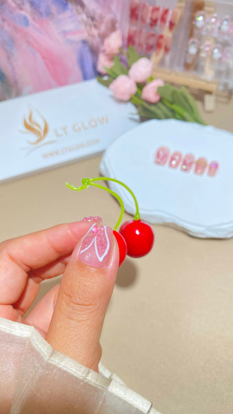 Hand showcasing a false nail design while holding a cherry, with an "LT Glow" backdrop and a set of nails on display in the background