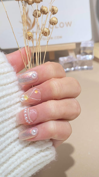 Hand adorned with handcrafted fake nails featuring delicate designs and embellishments, held against a backdrop with dried flowers and "LT Glow" sign