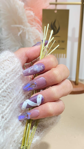 Hand showcasing intricately designed purple and white handmade press-on nails, holding delicate stems, with the LTGlow logo in the blurred background.
