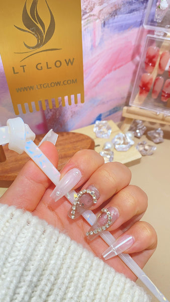 A person's hand displaying long, clear fake nails with delicate embellishments, held against a backdrop featuring LT GLOW branding
