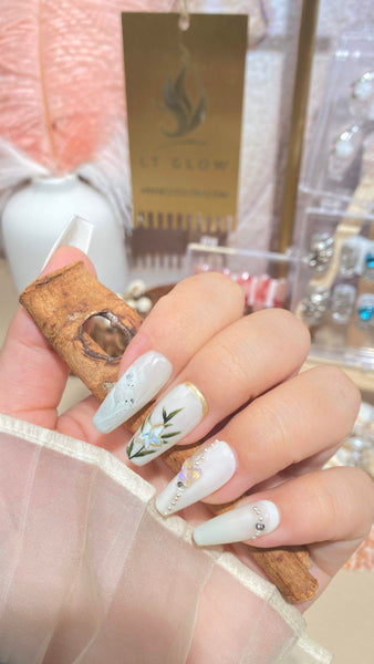 Elegant hand showcasing detailed fake nail designs with floral and gemstone accents, holding a rustic corked vial, set against a backdrop featuring the LTGlow brand sign