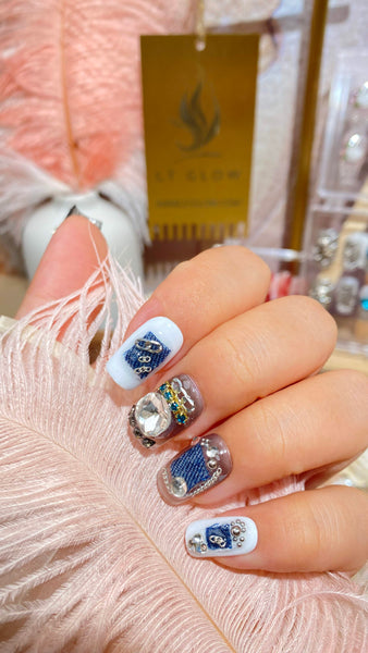 Handcrafted press-on nails with blue patterns and sparkling gemstone embellishments displayed against a pink feathery background