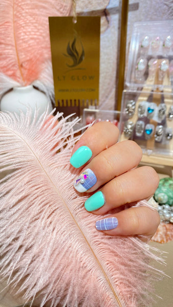 Hand showcasing vibrant acrylics nails in turquoise and pastel shades, with intricate designs, set against a plush pink background and LT Glow products on display