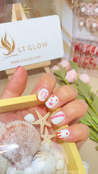 Close-up of a hand featuring handmade press-on nails with a summer-themed design, holding a golden frame, with the LT GLOW brand card and decorative seashells in the background