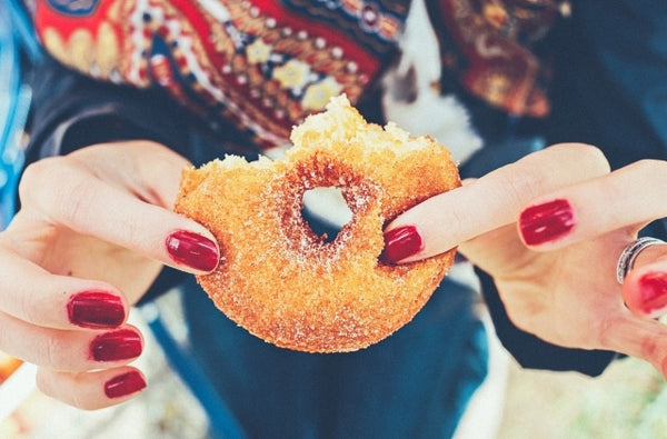 Close-up of hands with vibrant red handcraft fake nails holding a sugary donut, accentuating the bold nail color against the sweetness of the treat
