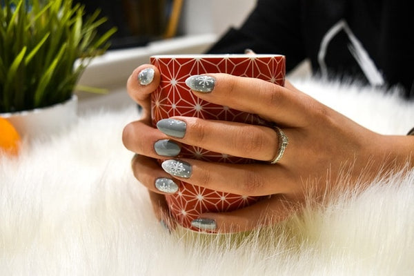 Elegant hands showcasing shimmering handmade false nails while holding a patterned red cup against a soft white fur backdrop